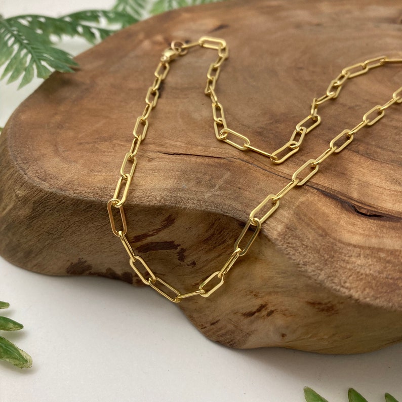 May include: A gold chain necklace with a paperclip chain style. The chain is laying on a wooden surface.