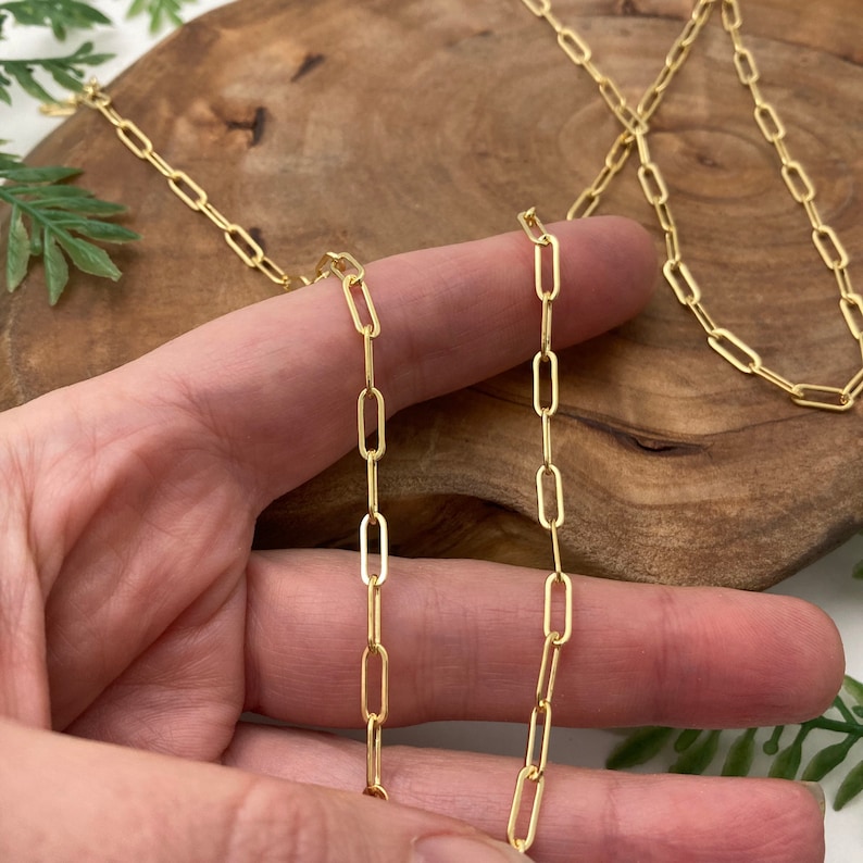 May include: A gold chain necklace with a delicate, oval-shaped link design. The chain is shown laid out on a wooden surface.