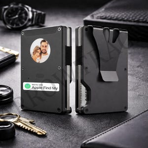 May include: A black metal wallet featuring a circular photo of a couple on the front, a white label with the text Works with Apple Find My, and a money clip on the back. The wallet has a slim, rectangular design with visible screws and side panels.