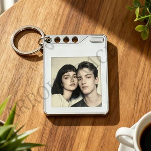 May include: A silver metal keyring with a rectangular photo frame. The frame contains a black and white photo of a couple. The keyring has a circular ring and small holes along the top edge.