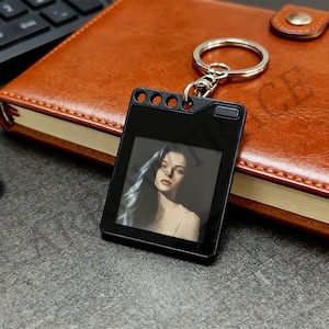 May include: A black rectangular keychain featuring a silver metal split ring and a central square insert displaying a grayscale photographic portrait of a person with long hair, set against a dark frame with three circular cutouts at the top.
