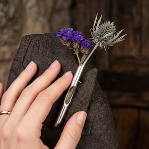 Op de afbeelding: Een zilveren reversbloemspeld met paarse en groene gedroogde bloemen. De speld is in de zak van een bruine blazer gestoken. Een gouden ring is zichtbaar aan een vinger.