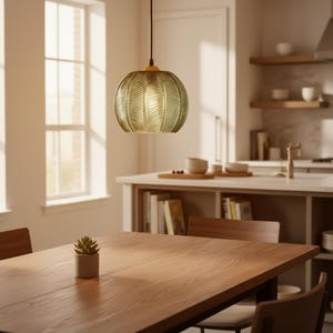 May include: A pendant light fixture with a textured, olive-green glass shade hangs above a wooden dining table. The light fixture has a gold-colored metal cap. A small potted succulent sits on the table. The room has a kitchen in the background.