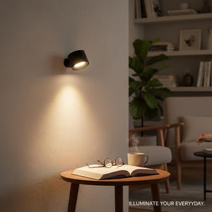 May include: A black, modern wall-mounted spotlight illuminates a warm, inviting glow. The light shines on a small, round wooden table with an open book, glasses, and a mug. The text "ILLUMINATE YOUR EVERYDAY" is at the bottom.
