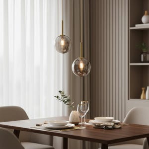 May include: Two pendant lights with spherical, smoky glass shades hang above a dining table. The lights have gold-coloured stems and emit a warm glow. The table is set with white dishes, glassware, and a vase with greenery, creating an elegant dining scene.