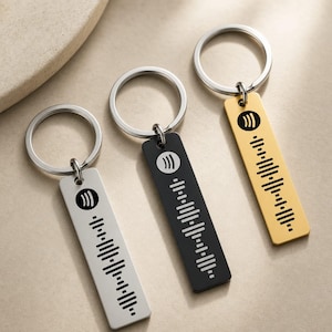 May include: Three rectangular metal keychains with silver-tone split rings. One is silver with black engraving, one is black with white engraving, and one is gold with black engraving. Each features a circular Spotify logo and a sound wave graphic.