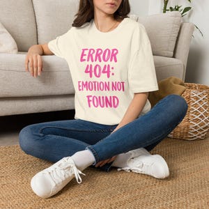 Op de afbeelding: Een crèmekleurig T-shirt met korte mouwen en de felroze tekst "ERROR 404: EMOTION NOT FOUND" op de voorkant. Het T-shirt wordt gecombineerd met een donkerblauwe spijkerbroek en witte sneakers voor een casual en eenvoudige outfit.