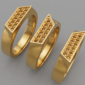 Puede incluir: Tres anillos dorados con una parte superior rectangular con múltiples pequeñas hendiduras circulares. Los anillos tienen un diseño geométrico con una banda plana y una parte superior ligeramente inclinada. Los anillos se muestran sobre una superficie gris.
