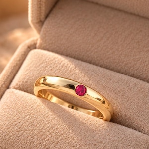 May include: A gold band ring featuring a single, small, round, bezel-set red gemstone centred on the top surface. The ring is displayed resting inside a beige, velvet-lined jewellery box with a hinged lid, showcasing a smooth, polished finish.