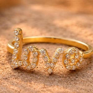 May include: A gold-toned ring featuring the word LOVE in cursive script, with the letters encrusted with small, clear, sparkling stones. The band is a simple, smooth gold-toned metal that completes the circular shape of the jewelry piece.