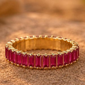 May include: A gold ring featuring a band of rectangular, deep pink gemstones. The ring rests on a textured, tan surface, with a blurred background of similar tones. The gemstones are closely set, creating a continuous line of colour.