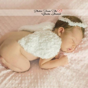May include: A newborn baby wearing a white crocheted angel wing costume and a white headband, sleeping on a pink blanket with a white polka dot pattern. The text "Photos From The Heart Christa Stewart" is visible above the baby.