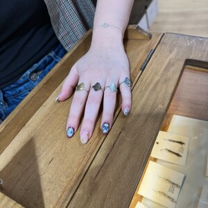 May include: A hand wearing four silver and gold rings with a ginkgo leaf design. The rings are on a wooden surface with other jewelry displayed in the background.