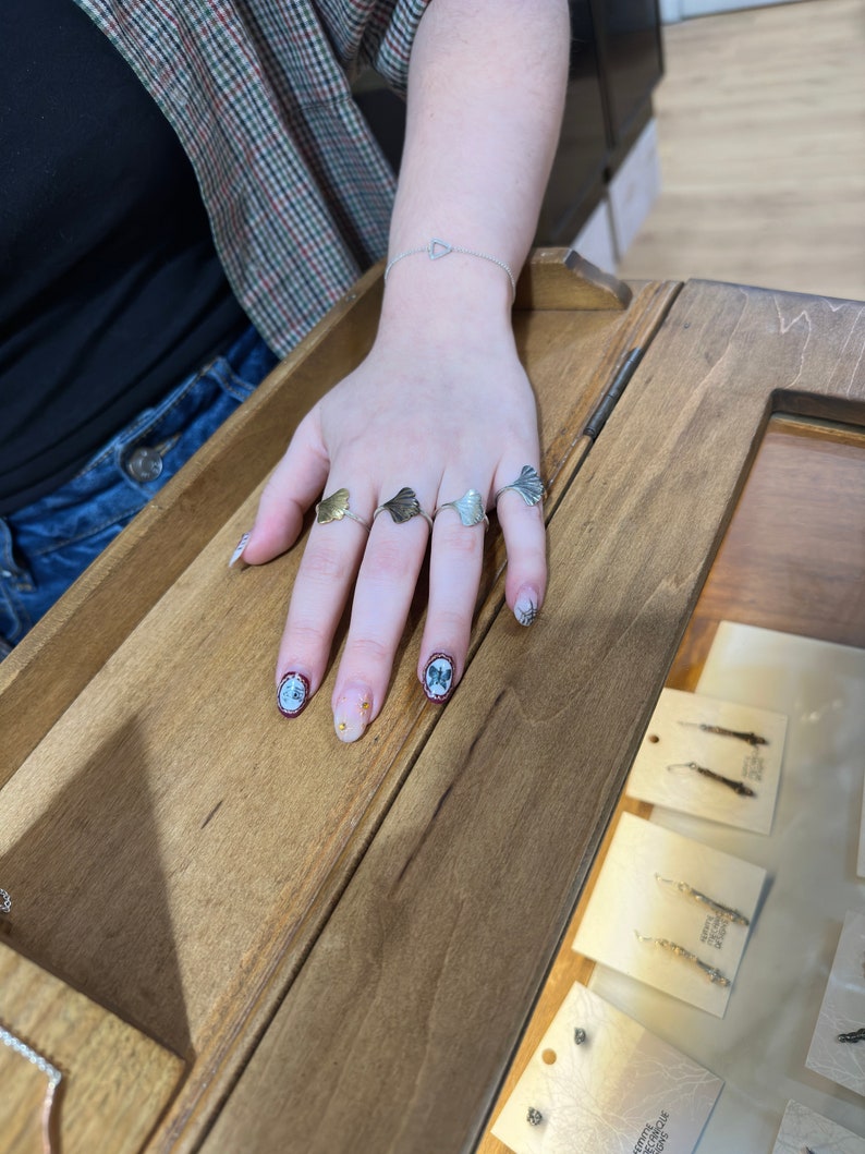 May include: A hand wearing four silver and gold rings with a ginkgo leaf design. The rings are on a wooden surface with other jewelry displayed in the background.