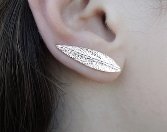 May include: A gold-colored leaf-shaped earring is worn on an ear. The earring has a textured surface, resembling the veins of a leaf. The earring is a unique piece of jewelry.