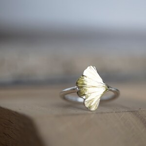 May include: A silver ring with a gold ginkgo leaf design. The ring is sitting on a wooden surface.