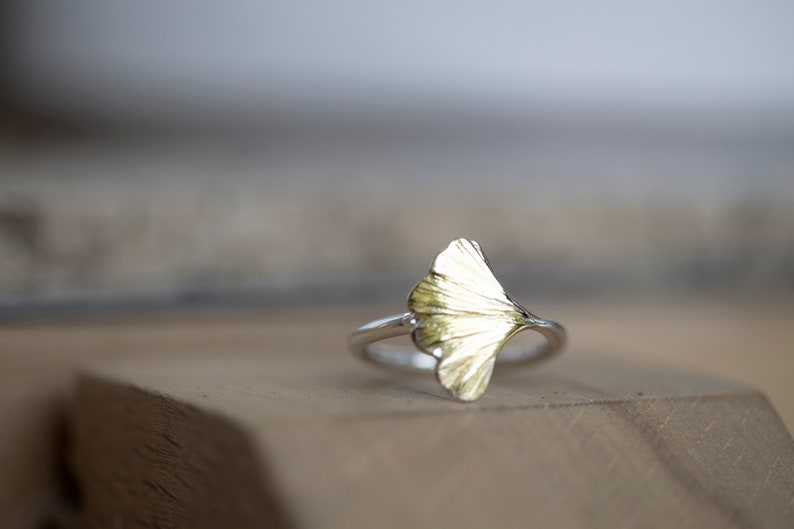 May include: A silver ring with a gold ginkgo leaf design. The ring is sitting on a wooden surface.