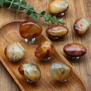 May include: A collection of polished agate stone cabinet knobs in shades of brown, orange, and cream. Each knob is mounted on a silver-colored base. The knobs are arranged on a wooden tray and surface.