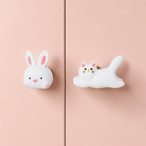 May include: White bunny and cat cabinet knobs. The bunny knob has pink inner ears and a small pink nose. The cat knob is lying down with a tail up and has black whiskers and pink ears. Both knobs are on a pink cabinet door.