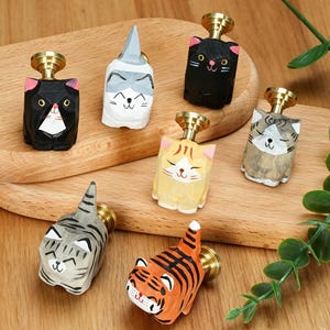 May include: A collection of six hand-painted wooden cat-shaped cabinet knobs, each featuring a unique feline pattern and color scheme, including black, white, orange, and gray tabby designs, all topped with a polished brass metal mounting base.