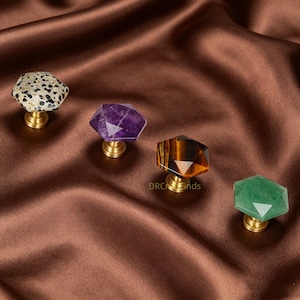 May include: Four gemstone cabinet knobs with brass bases. The gemstones are in various colours and patterns, including a speckled white and black, purple, brown, and green. The knobs are set against a brown satin background.