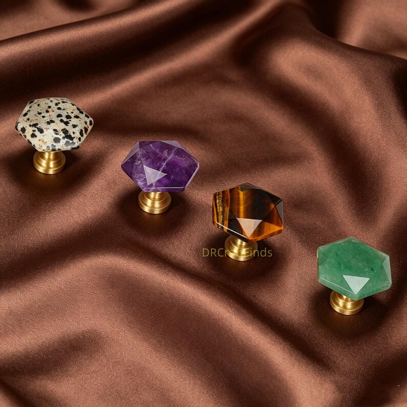 May include: Four gemstone cabinet knobs with brass bases. The gemstones are in various colours and patterns, including a speckled white and black, purple, brown, and green. The knobs are set against a brown satin background.