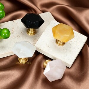 May include: Four faceted gemstone cabinet knobs in black, yellow, white, and pink. Each knob is mounted on a brass base. The knobs are displayed on square, off-white stone tiles against a brown satin background. The text "DRC Finds" is visible.