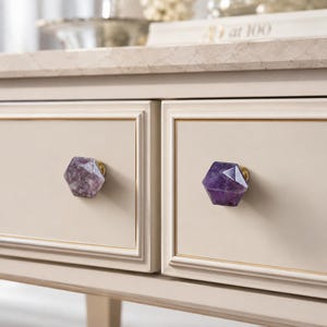 May include: Close-up of a cream-coloured dresser with two drawers. Each drawer features a faceted, purple amethyst knob with a gold-coloured base. The dresser has a light-coloured marble top and gold trim.