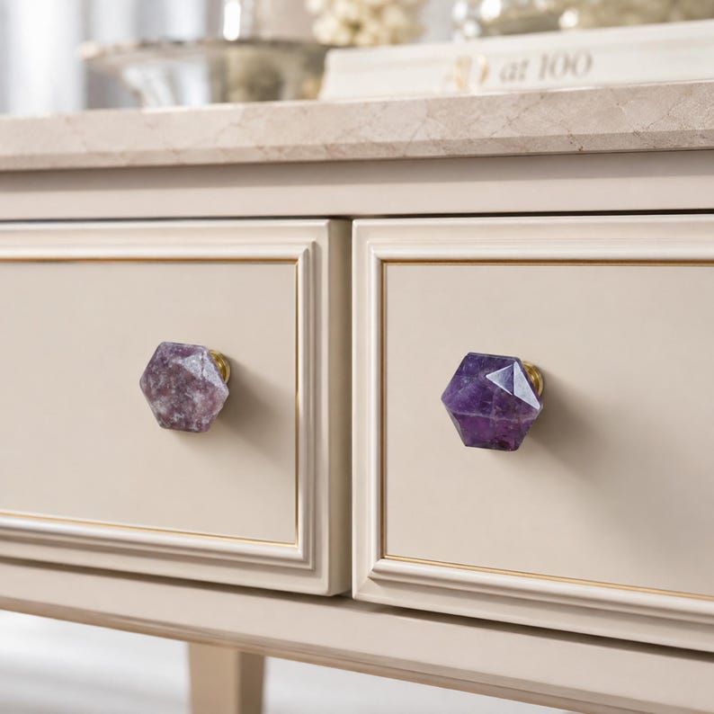May include: Close-up of a cream-coloured dresser with two drawers. Each drawer features a faceted, purple amethyst knob with a gold-coloured base. The dresser has a light-coloured marble top and gold trim.