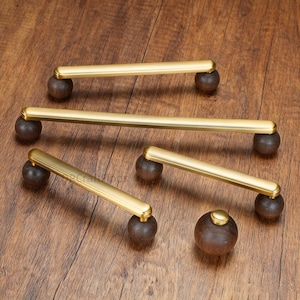 May include: A collection of brass-toned cabinet pulls with dark brown wooden ball accents. The pulls are rectangular with rounded edges. One round wooden knob is also present. The hardware is displayed on a wooden surface.