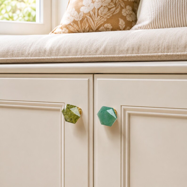 May include: Two cabinet knobs with faceted gemstone designs. One is green with gold hardware, the other is a lighter green with gold hardware. The knobs are mounted on a cream-coloured cabinet door.