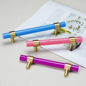 May include: Three cylindrical cabinet pulls with gold-toned metal mounts. The top pull is bright blue, the middle pull is pink with the text DRCraftFinds, and the bottom pull is purple. Each handle features two metallic supports for attachment.