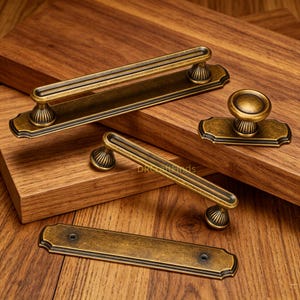 Op de afbeelding: Een set antiek messingkleurig deurbeslag bestaande uit een lange ladehandgreep met rechthoekige achterplaat, een kleinere handgreep, een ronde meubelknop op een rechthoekige basis en een losse rechthoekige plaat met twee schroefgaten.