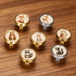 May include: A set of seven round cabinet knobs featuring watercolor illustrations of woodland animals including a fox, deer, bear, rabbit, owl, raccoon, and skunk. The knobs have either a brushed gold or silver metal finish with a cream center.