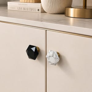 May include: A pair of cabinet knobs, one black and faceted, the other white with grey marbling. Both have gold-coloured bases and are attached to a light beige cabinet door. The knobs are hexagonal in shape. The cabinet door has a gold trim.