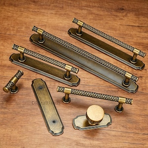 Könnte beinhalten: Eine Auswahl an antiken bronzefarbenen Schrankbeschlägen, darunter Griffe, Züge und ein Knauf. Die Griffe weisen ein strukturiertes, gewebtes Design auf. Die Hardware wird auf einer Holzoberfläche präsentiert.