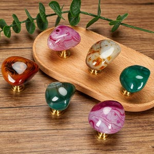 Puede incluir: Un conjunto de seis tiradores de armario de piedras preciosas en varios colores, incluyendo rosa, verde, naranja y blanco. Cada tirador está montado sobre una base dorada y se exhibe sobre una bandeja y mesa de madera.