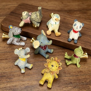 Puede incluir: Colección de figuras de animales caprichosas, incluyendo un oso, un elefante, un pato, un panda, una rana y un león. Cada figura está pintada con colores brillantes y tiene una bocina dorada. Sobre una superficie de madera.