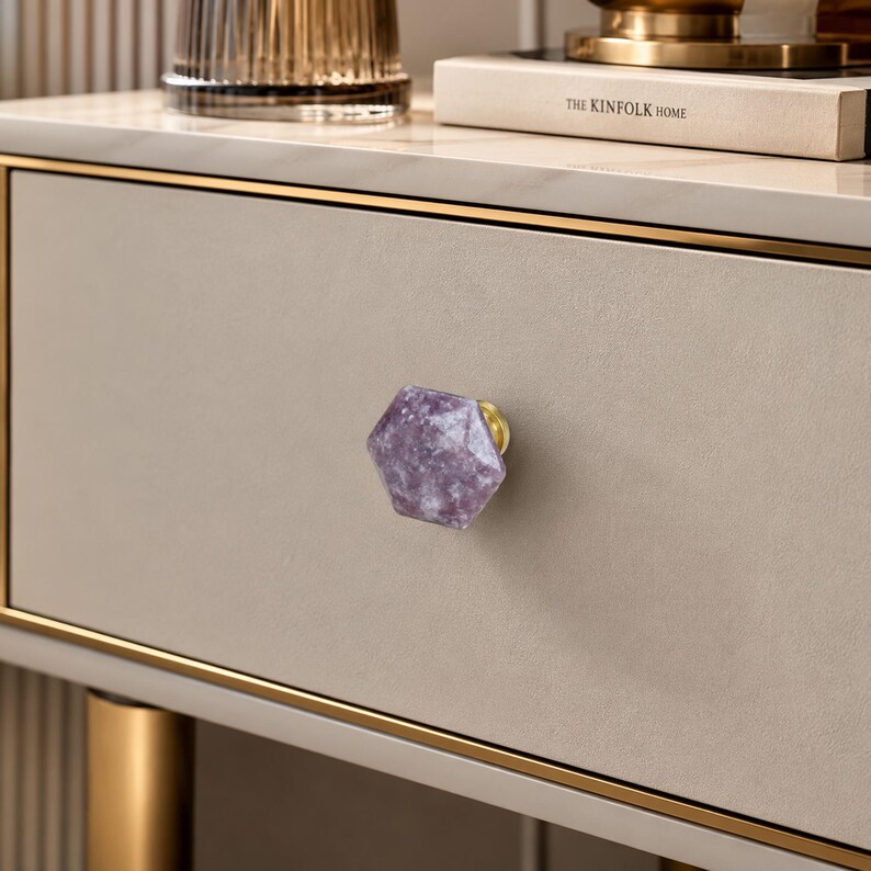 May include: A light beige drawer with a hexagonal purple and white knob. The drawer has gold trim. The knob has a gold base. The Kinfolk Home book is visible in the background. The knob is a decorative element.