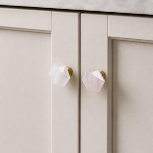 May include: Close-up of two cabinet doors with decorative knobs. One knob is white, the other is pink, both with a faceted, geometric design. The knobs have a gold-coloured base. The cabinet doors are a light beige colour.