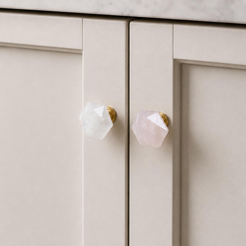 May include: Close-up of two cabinet doors with decorative knobs. One knob is white, the other is pink, both with a faceted, geometric design. The knobs have a gold-coloured base. The cabinet doors are a light beige colour.