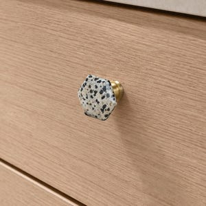 May include: A close-up of a hexagonal cabinet knob with a speckled, off-white and black design. The knob has a brass base and is mounted on a light-coloured wooden drawer front. The knob adds a decorative touch to furniture.