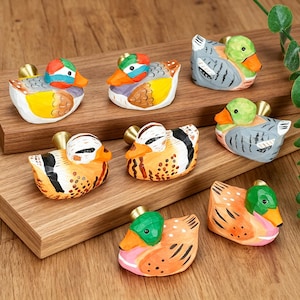 Peut inclure: Une collection de sept boutons de tiroir en forme de canard sculptés à la main, chacun présentant un motif peint unique avec des nuances de vert, orange, jaune, gris et noir, dotés d'une tige de fixation en métal de couleur laiton à l'arrière.