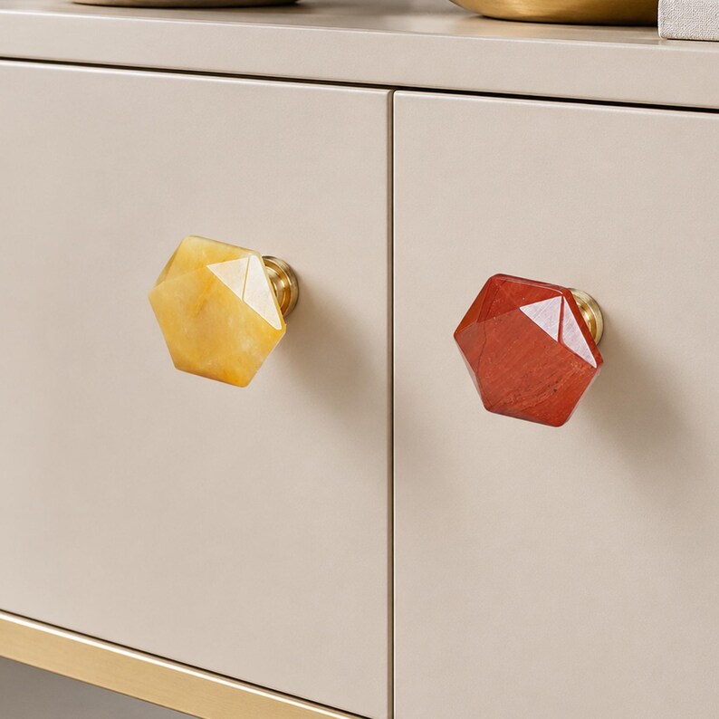 May include: Close-up of two cabinet knobs. One is yellow, the other is red, both with a faceted, geometric design. The knobs are mounted on a light beige cabinet door with gold hardware.