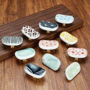 May include: A collection of ceramic cabinet knobs with various patterns and colors. The knobs are white with designs in black, brown, red, yellow, blue, and green. Each knob has a brass base and is displayed on a wooden surface.