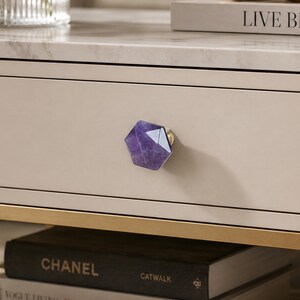 May include: A close-up of a drawer with a purple amethyst crystal knob. The knob is a faceted, geometric shape with gold hardware. The drawer is a light beige colour, and the surface above is marble.