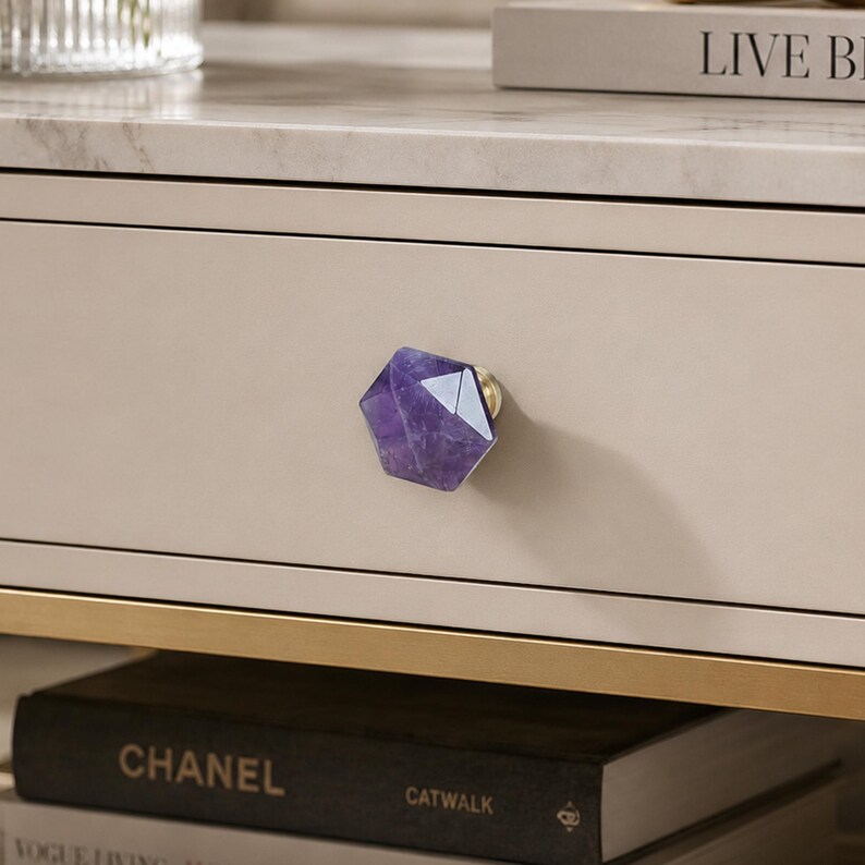 May include: A close-up of a drawer with a purple amethyst crystal knob. The knob is a faceted, geometric shape with gold hardware. The drawer is a light beige colour, and the surface above is marble.
