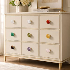 May include: A cream-coloured chest of drawers with nine drawers, each featuring a unique gemstone knob in various colours. The knobs include white, grey, red, green, yellow, and purple. The chest has gold trim and legs.