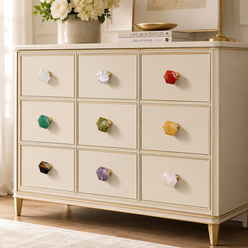 May include: A cream-coloured chest of drawers with nine drawers, each featuring a unique gemstone knob in various colours. The knobs include white, grey, red, green, yellow, and purple. The chest has gold trim and legs.