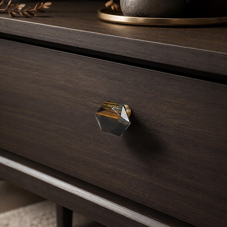 May include: A close-up shot of a dark brown wooden drawer with a hexagonal knob. The knob has a gold base and a clear, amber-toned crystal. The drawer is part of a larger piece of furniture, possibly a chest of drawers or bedside table.
